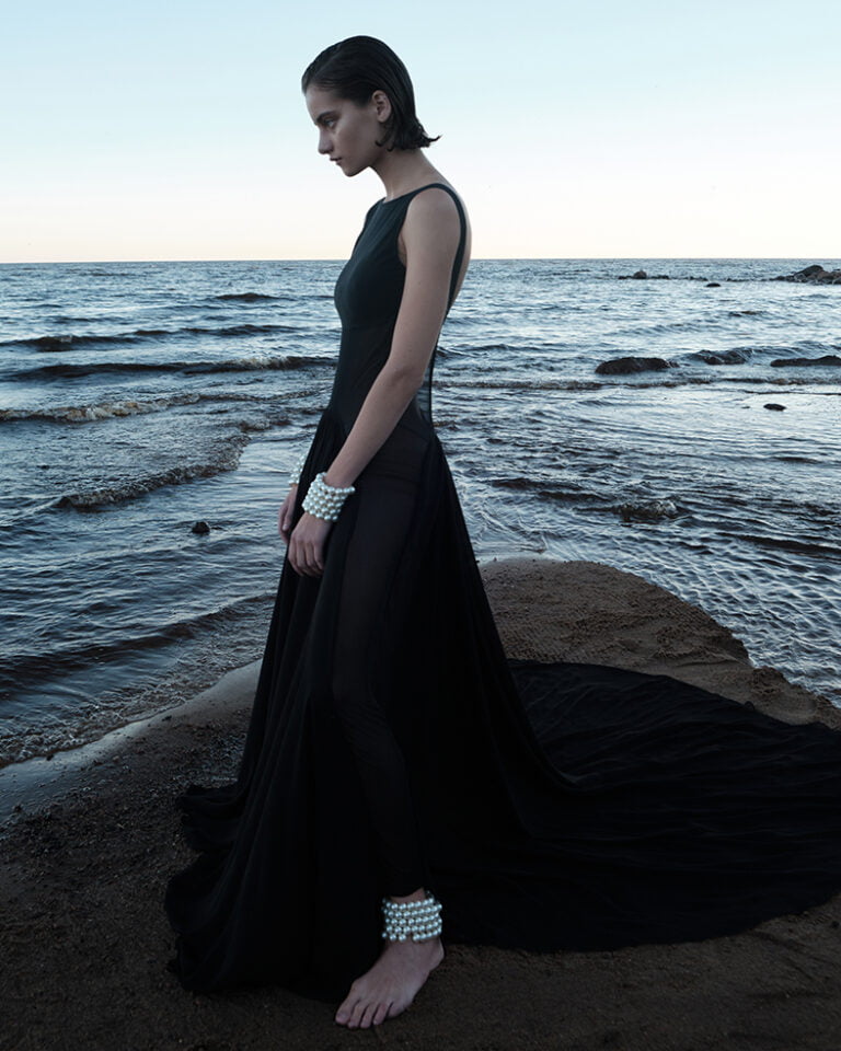 Photograph of a model in long black dress with big pearl bracelets on her arms and legs