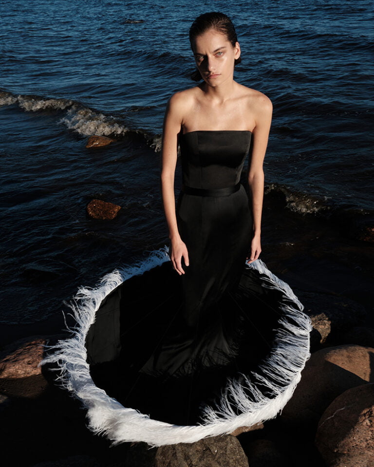 Photograph of a fashion model in black skinny dress with white feathers at the bottom.