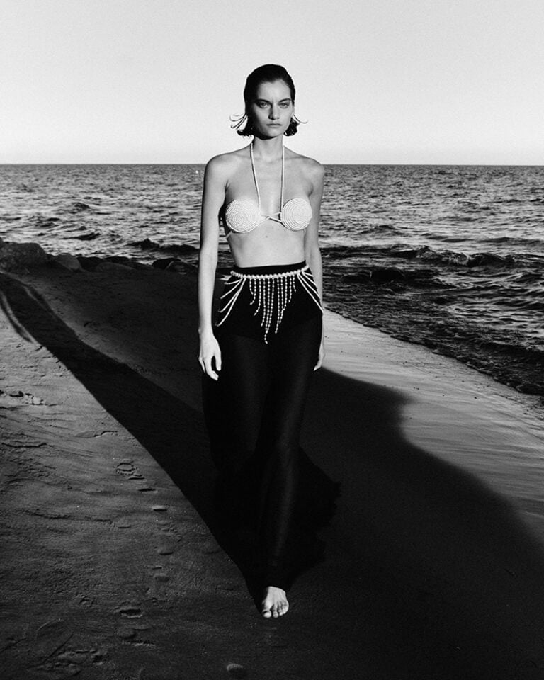 A model wearing a long black skirt and pearls is walking along the beach