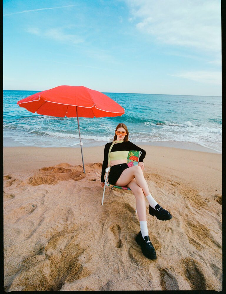 A model sitting at the beach next to a red umbrella. Sport fashion photography.