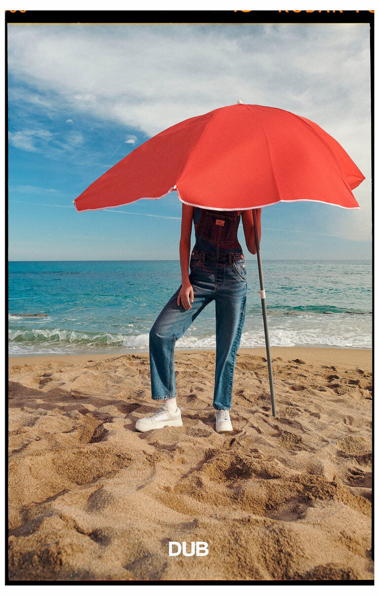 Photograph of a model at the beach, her head is hidden under a red umbrella.