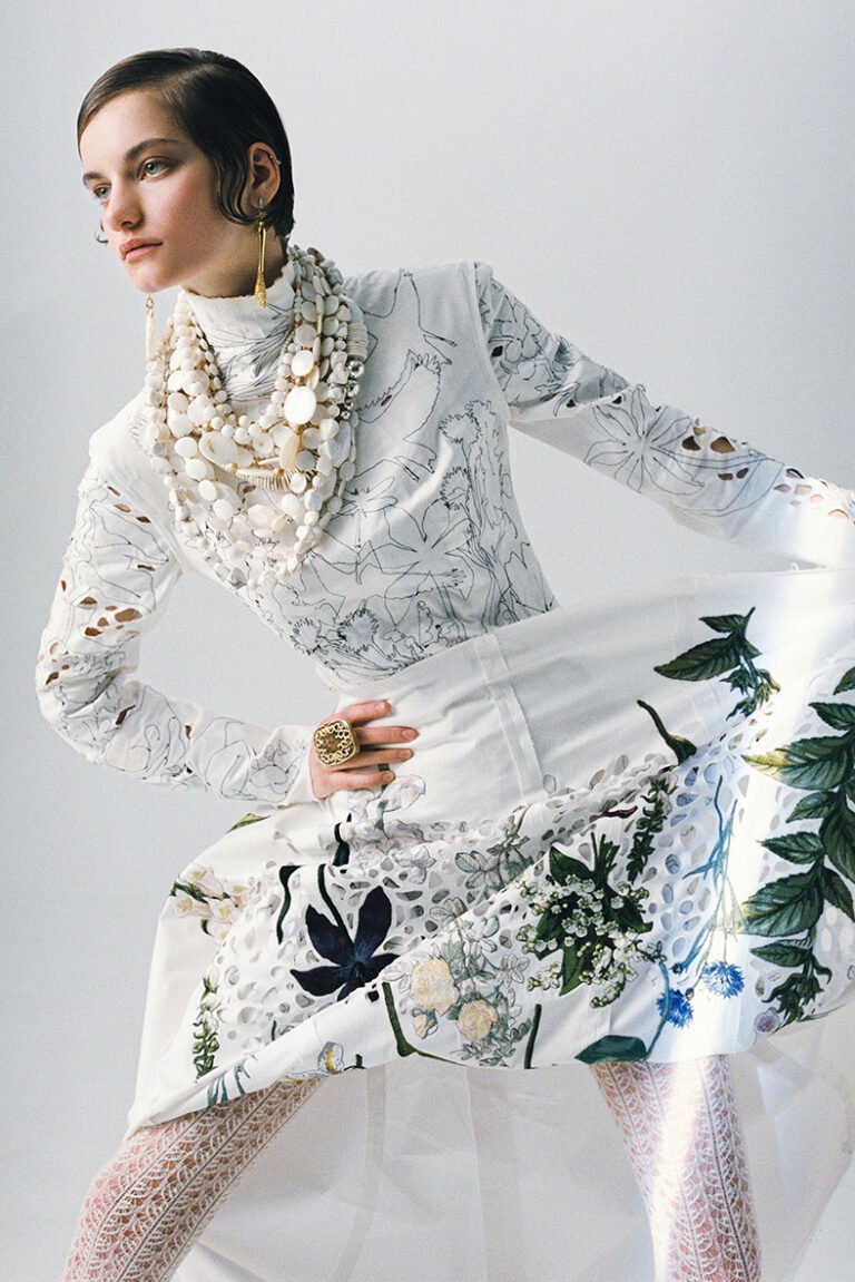 Model in a white dress with beads on her neck, earrings in her ears and a big finger ring